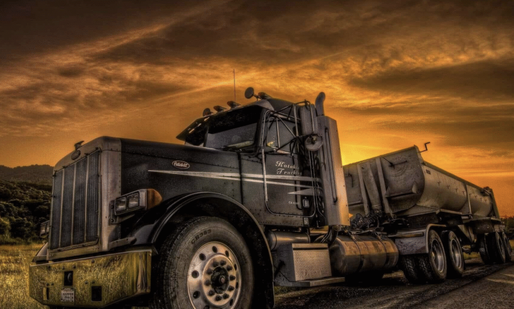 Low-angle photograph of a heavy-duty semi-truck and its trailer, set against a stunning, intensely coloured sky, likely at sunrise or sunset.