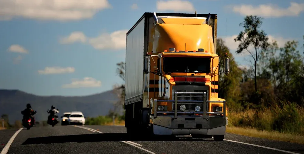 A large semi-trailer truck traveling on a paved highway, with other vehicles in the distance, set against a bright, sunny, and rural landscape.